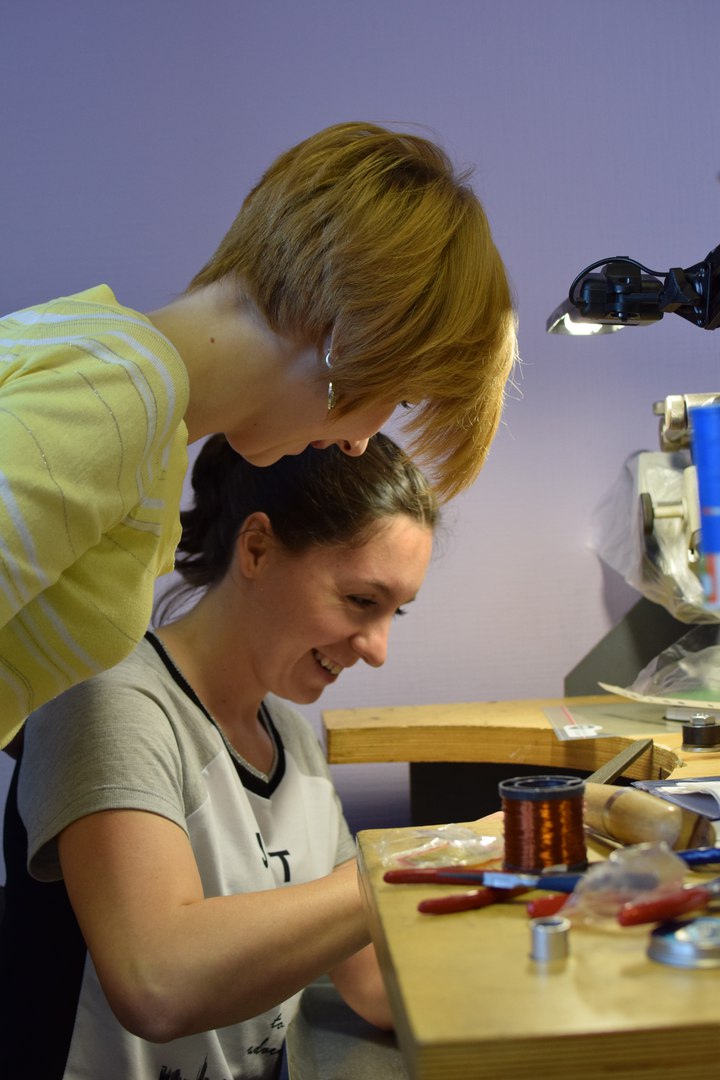 In the photo, Yulia Pavlenko sits at a table crafting a pendant. The image captures a workshop class. Next to Yulia stands her instructor, observing her work. Both are smiling. The wall is purple, illuminated by warm yellow light. The workbench, made of light-colored wood, holds various tools