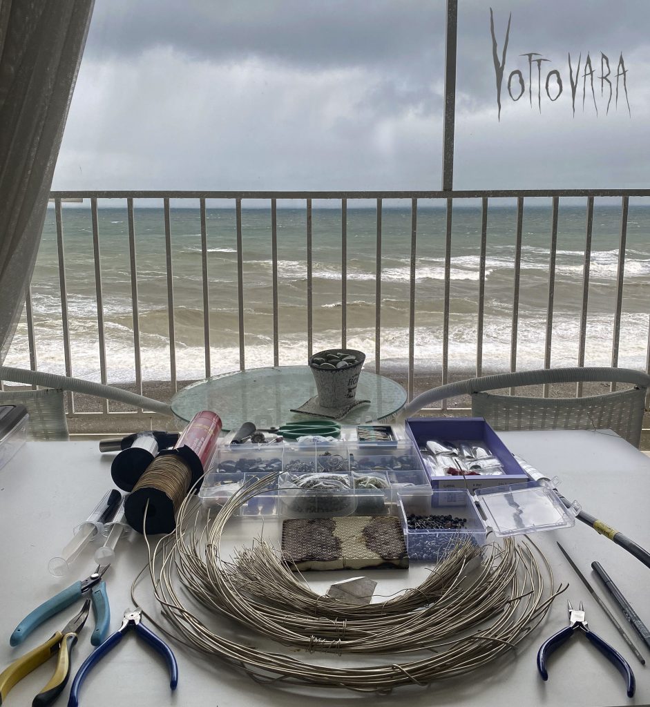 A jewelry artist's workspace featuring tools, wire, and crafting materials on a table by a window overlooking the sea. The waves and cloudy sky in the background create a peaceful and inspiring atmosphere.