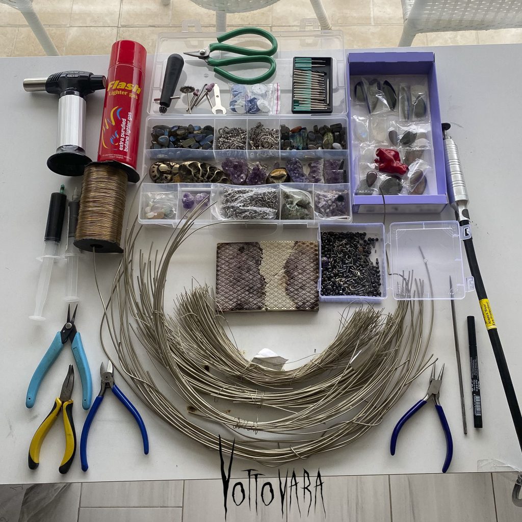 A workspace featuring jewelry-making tools, spools of wire, a torch, and organized boxes of semi-precious gemstones and findings. These are the materials and tools used to create Vottovara artifacts.