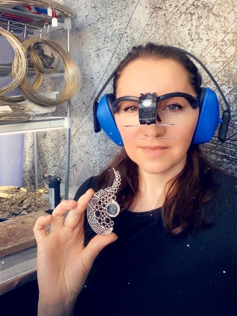 Yulia Pavlenko, wearing magnifying glasses and ear protection, holds a handcrafted wire-woven pendant with a central stone in her workshop, surrounded by tools and materials.